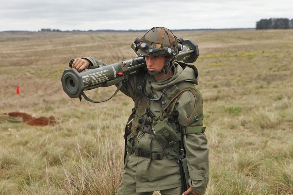 Militar da FOROP/CAAdEx, em seu uniforme peculiar e trajando o simulador pessoal BT47, conduz o simulador do AT4 para sua posição de tiro